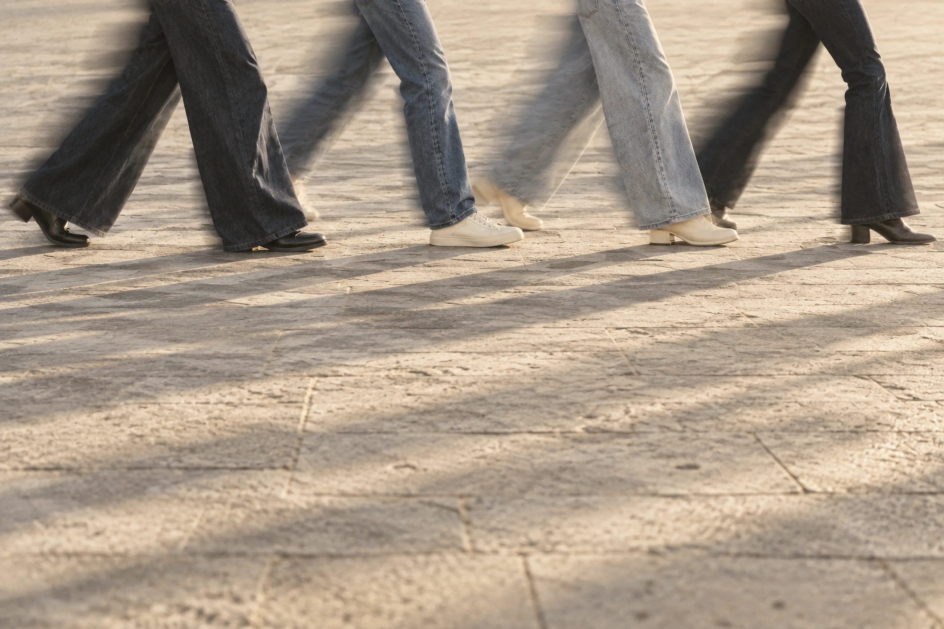 Four people walking across sunlit paving in wide-leg jeans in different washes, their legs blurred mid-stride, long shadows on the stone.